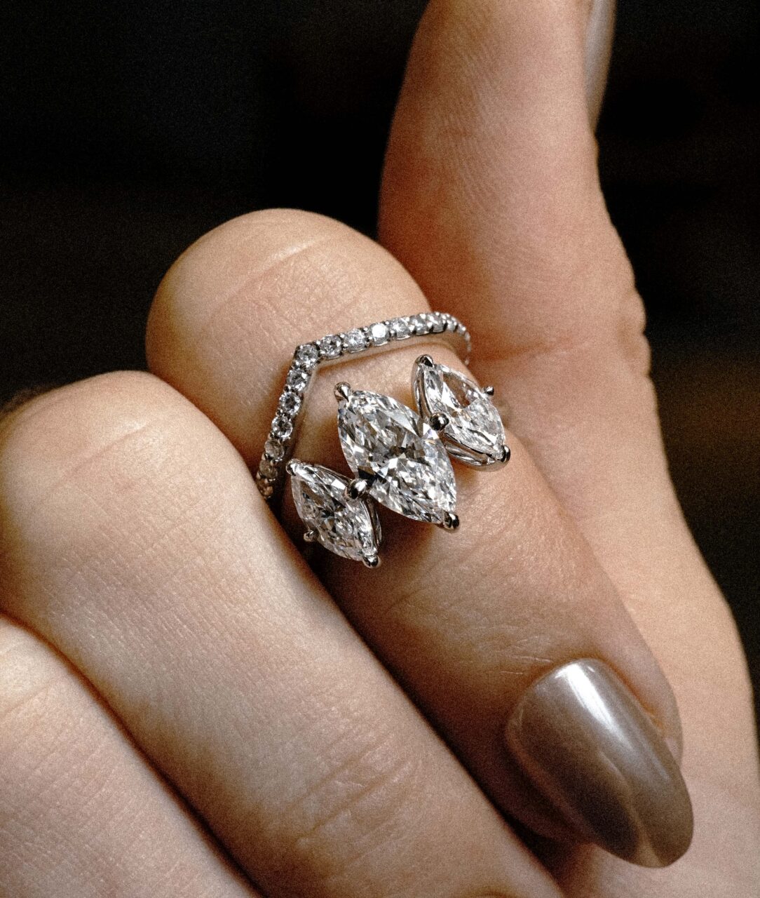 A three stone engagement ring has three Marquise cut diamonds. Below the ring is a diamond pave chevron band.
