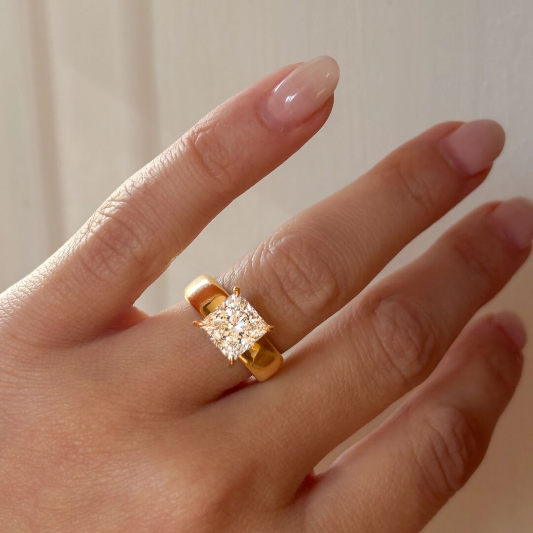 A hand with neutral pained nails wear a princess cut, prong set engagement ring with a chunky, 18K gold band 