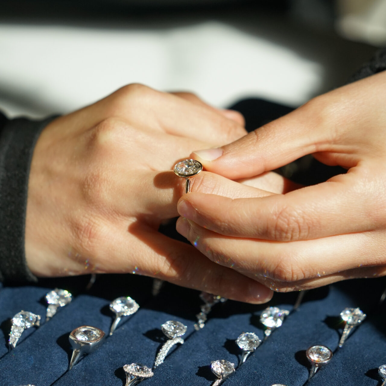 A person tries on an oval cut solitaire engagement ring, below their hand is a blue velvet tray filled with diamond engagement rings 