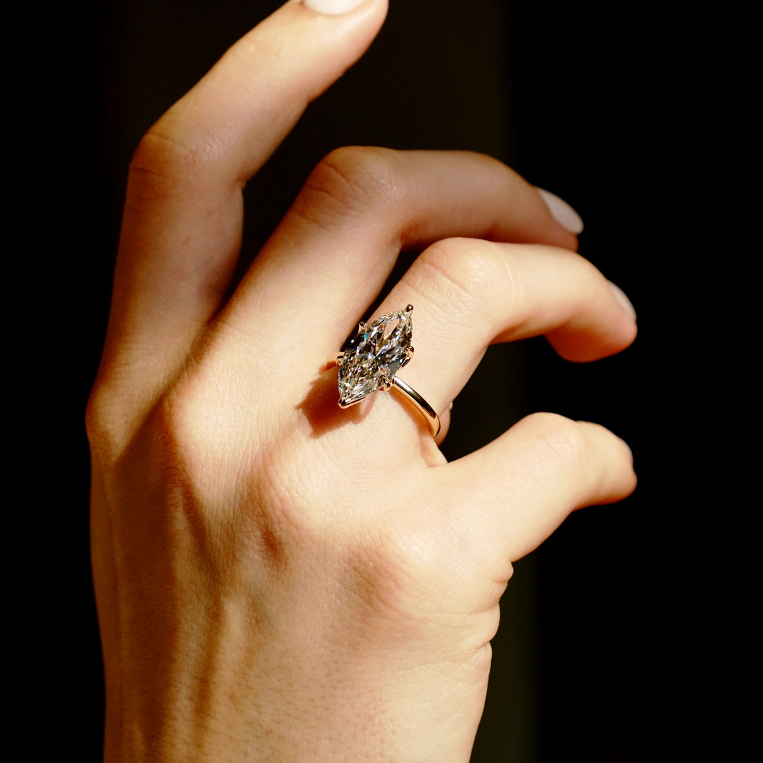 A six prong Marquise diamond solitaire engagement ring inyellow gold is worn against a black backdrop