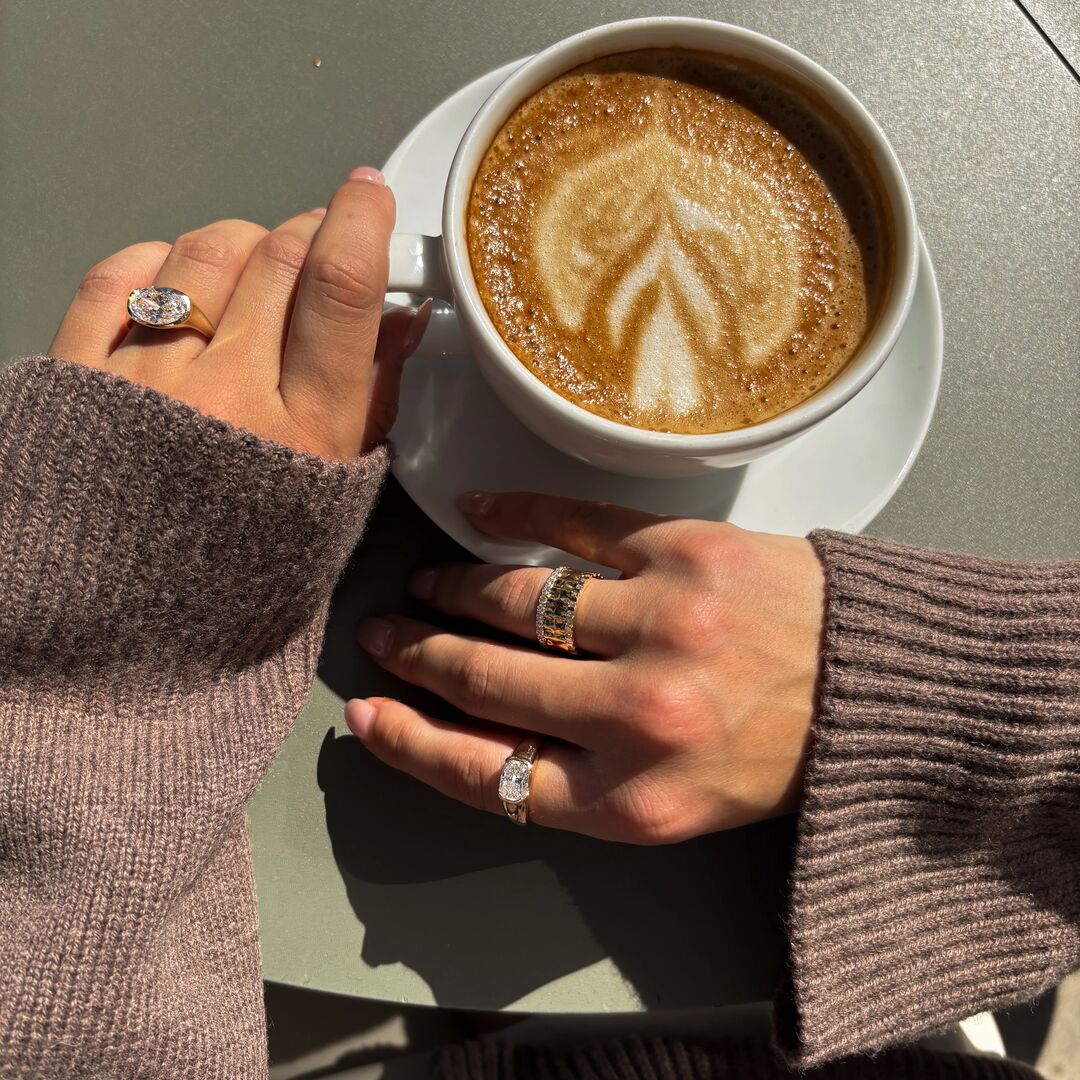 A woman in a chunky sweater holds a cup of coffee and wears a variety of diamond jewelry 