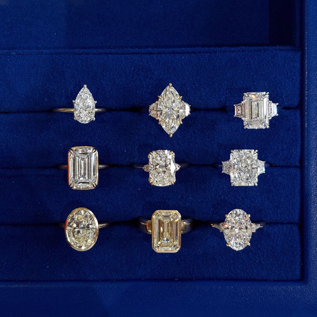 Six rings on display in a blue velvet box. Top row: A two tone, pear cut engagement ring, a three stone marquise cut ring, and a three stone emerald and trapezoid engagement ring. Second row: A collet set emerald cut solitaire, a three stone, elongated cushion and trillion ring, and a three stone radiant and trapezoid engagement ring. Third Row: a bezel set, oval cut solitaire, a collet set emerald cut diamond on a chunky yellow gold band, and a three stone, oval and tapered baguette ring 